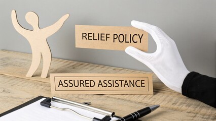Hand Holding Card with Text on Relief Policy and Assured Assistance beside Wooden Figure on Table in Office Setting