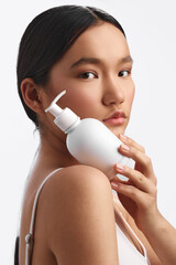 asian woman holds a lotion bottle near her shoulder in a clean studio, close up portrait with soft...