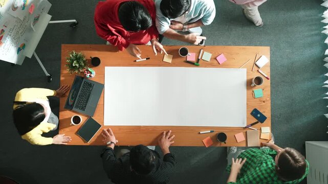 Top view of diverse business team brainstorming marketing idea while project manager standing at whiteboard. Multicultural people discuss strategy while standing around at meeting table. Convocation.