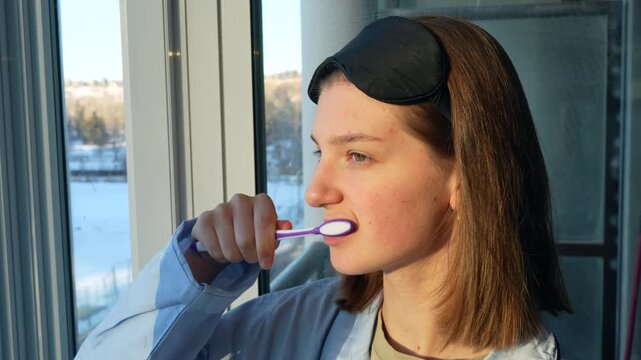 Young woman doing usual morning routine - brushes teeth looking at the morning city view smiles and gets ready for a new day