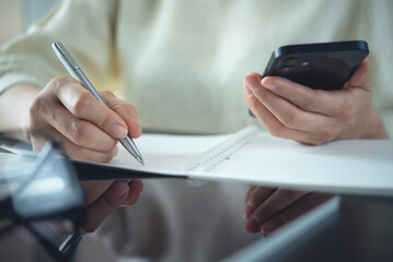  Business woman holding smartphone in hands, writing notes in personal daily planner, planning workday, checking schedule and handwriting  information at home office, job scheduling