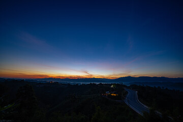 A scenic winding road stretches through lush green hills and layered mountains under a warm sunrise, creating a peaceful and inspiring countryside landscape. Perfect for travel, adventure, and journey