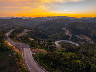 Aerial view of A winding road twisting across tall mountain peaks in Nan Province, illuminated by soft sunset glow and surrounded by a peaceful natural landscape.