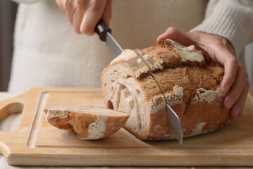 Close-up of a woman in a white cozy sweater slicing sourdough bread on a wooden board