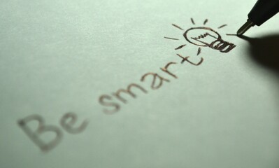 A hand drawing a lightbulb with the words Be smart written on a white paper from a close-up viewpoint