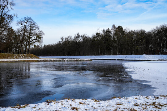 Sunny winter landscape with a partially frozen body of water, surrounded by snow-covered shores and a forest area in the background. Peaceful atmosphere in the Nymphenburg Palace Park in Munich
