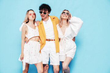 Fototapeta premium Group of young three stylish friends posing in studio. Fashion man and two cute female dressed in casual summer clothes. Smiling models having fun. Cheerful women and guy near blue wall, sunglasses