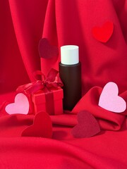 A beautiful festive arrangement featuring a gift box with a bow and a bottle of cosmetics against a backdrop of red draped fabric and hearts symbolizing love. Happy birthday or Valentine's Day greetin
