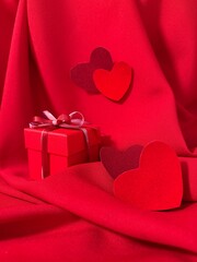 A beautiful arrangement in red and scarlet tones with a Valentine's Day gift box against a backdrop of draped red fabric and Valentine hearts symbolizing love. Happy Valentine's Day concept