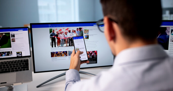 Man Reading Multiple Online News Articles On Mobile Phone And Laptop