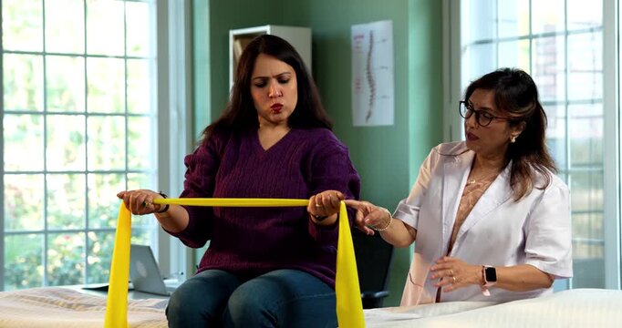 Indian lady patient with physiotherapist performing triceps and shoulder strengthening exercises using resistance band in modern clinic under senior guidance for rehabilitation and recovery