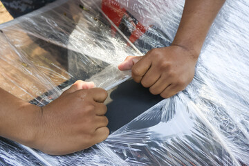 Closeup hands tightly stretching clear plastic wrap over dark item for protective shipping with careful focused preparation and diligent manual labor showing secure packing process