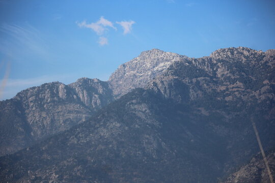Close-up view of a rugged granite mountain face with patches of snow and scattered evergreen trees under a clear sky. - Powered by Adobe
