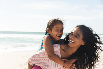 Fototapeta premium Cheerful woman giving piggyback ride to daughter at beach against sky with copy space