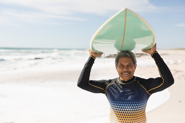 Portrait of smiling senior woman wearing wetsuit carrying surfboard on head at sunny beach