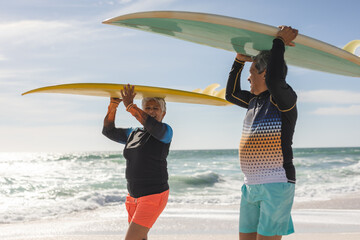 Obraz premium Happy multiracial senior couple looking at each other while carrying surfboards on heads at beach