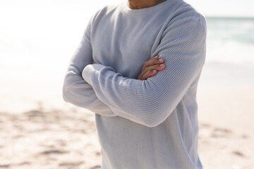 Midsection of retired senior man standing with arms crossed at beach during sunny day