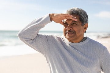 Retired senior man shielding eyes while looking away at beach during sunny day