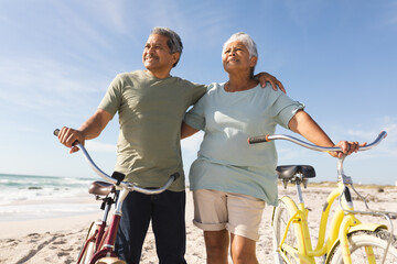 Naklejka premium Smiling senior multiracial couple looking away with bikes at beach against sky during sunny day