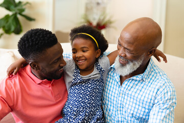 African american grandfather, father and granddaughter smiling looking at each other at home