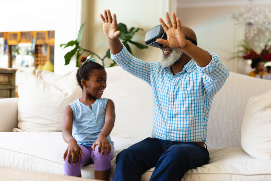 African american girl smiling and her grandfather wearing vr headset gesturing sitting on the couch - Powered by Adobe