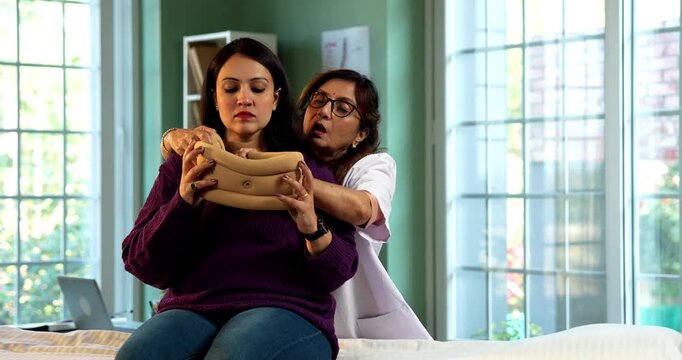 Indian lady physiotherapist treating neck pain of young female patient while fitting cervical support collar in modern clinic, both showing ok gesture and confidence in recovery care