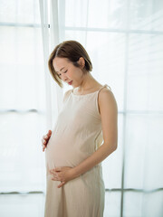Happy Asian pregnant woman lovingly touching her unborn baby while standing by the window at home.