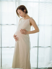 Happy Asian pregnant woman lovingly touching her unborn baby while standing by the window at home.