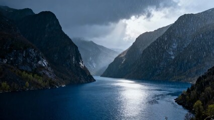 A serene mountain lake surrounded by steep cliffs under a dramatic, cloudy sky with sunlight breaking through.