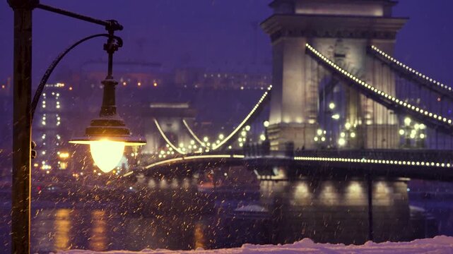 Budapest, Hungary - January 6, 2026: Snowflakes fall past a glowing street lamp, illuminating the dark waters of the Danube and the iconic lit arches of the Chain Bridge in Budapest.