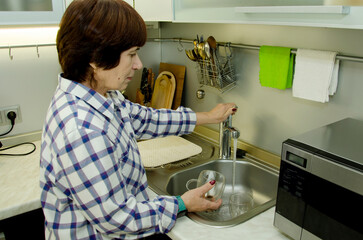 Elderly woman washing glass mug under running tap in modern kitchen, focused expression, domestic routine, quiet responsibility, home maintenance, everyday care, simple task.