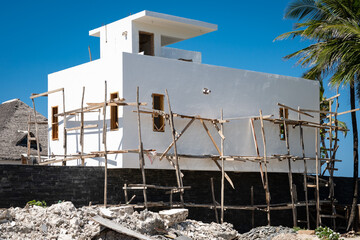 A modern white concrete house under construction in Zanzibar, featuring traditional wooden scaffolding against a clear blue sky and tropical palm trees.