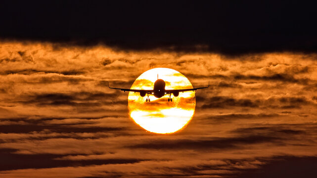View of a silhouette airplane ascending against a fiery sunset, its wings outstretched, creating a striking contrast against the glowing sun, Schiphol, Noord-Holland, Netherlands.
