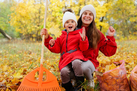 A mother and her child smile while raking leaves in a park filled with trees showing autumn colors. They have a bag full of leaves beside them - Powered by Adobe