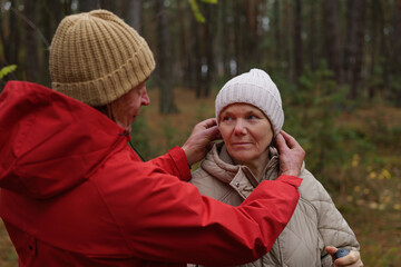 Two people are in a forest. A man adjusts a woman's hat. They wear warm clothing. The trees show autumn colors. It is late afternoon with soft sunlight