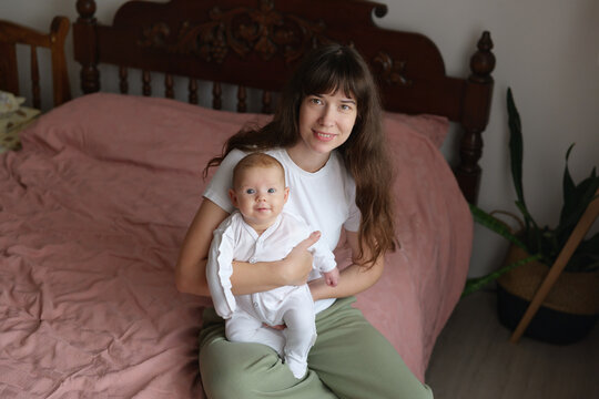 Mother sits on bed with baby, both smiling. The room has soft light and a simple, warm atmosphere. They seem to enjoy their time together
