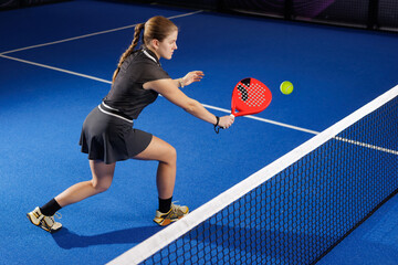 Focused female padel player hits volley at the net on indoor blue court