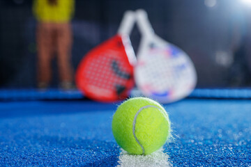 Close up padel ball on blue indoor court with rackets and net in background