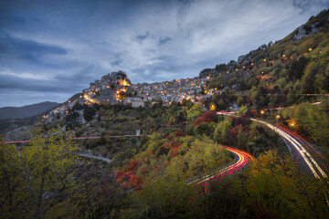 View of a medieval village perched atop a hill, illuminated by warm lights contrasting with the cool evening sky, with winding roads below traced by streaks of car lights, Subiaco, Lazio, Italy.