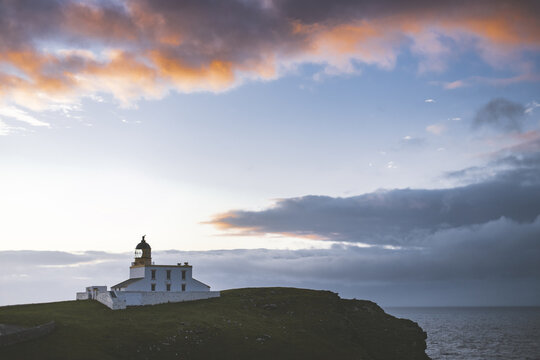 View of a white lighthouse standing proudly on a verdant cliff under a sky ablaze with the fiery hues of sunset and the dark clouds, Isle of Skye, United Kingdom.