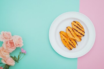Slices of grilled sweet potato on a white plate with pink roses on a pastel pink and teal background