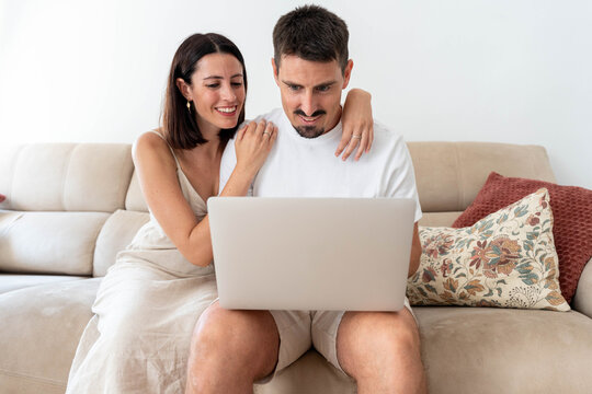 Couple enjoying leisure time at home, relaxing on the sofa, smiling while looking at a laptop computer together, interacting with online content and searching on internet - Powered by Adobe