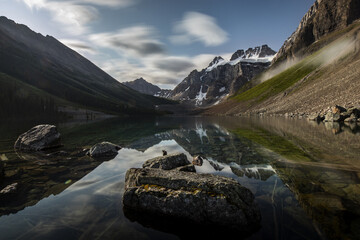 View of the still, reflective lake mirrors the rugged, snow-capped mountains under a sky streaked with wispy clouds, Banff, Alberta, Canada.
