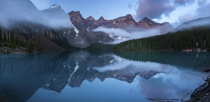 View of serene turquoise waters mirroring jagged, snow-dusted peaks and emerald forests under a sky veiled in soft, ethereal clouds, Banff, Alberta, Canada.