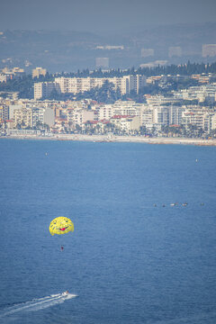 View of a smiley-faced parasail soars above the azure sea, the city skyline fading into the misty horizon, a speedboat cutting through the water, Nice, France.