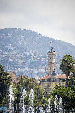 View of fountains dancing in the foreground, contrasting with the clock tower, and rolling hills under a soft sky, creating a serene cityscape, Nice, France.