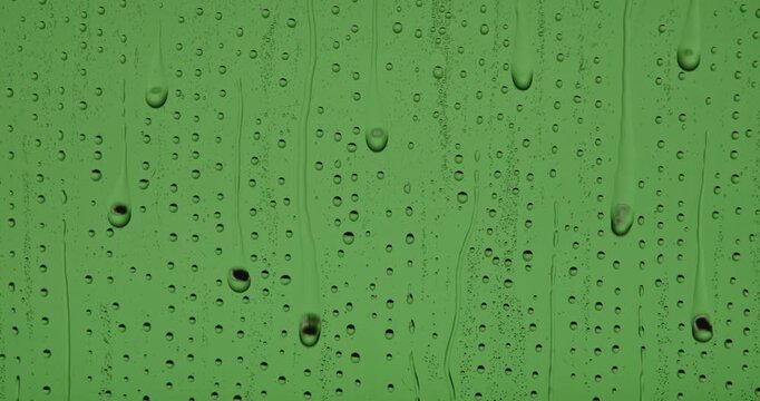 Close up of wet glass with dripping water drops and streaks of rain, on a bright green chroma key background, perfect for visual effects and background replacement. Slow motion.