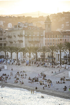 View of sun-kissed sands meet the turquoise sea as people gather along the beach, framed by elegant buildings and swaying palms, Nice, France.