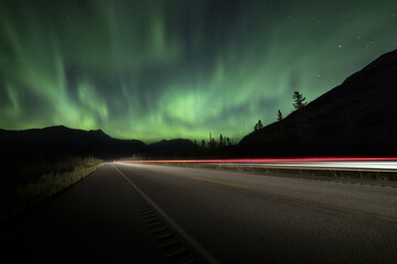 View of vibrant green aurora borealis dances above the silhouetted mountains, contrasting with the blurred red and white lights of passing vehicles on the dark asphalt road, Jasper, Alberta, Canada.