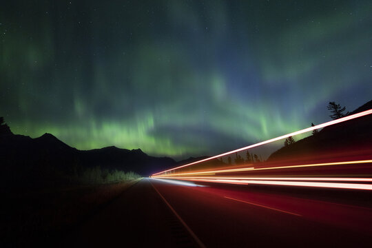 View of vibrant aurora borealis dances above the dark silhouettes of mountains, as car lights streak across the highway, Jasper, Alberta, Canada.